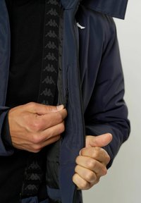 Hände, die die Innentasche einer marineblauen Jacke über einem schwarzen Hemd mit einem gemusterten Trageriemen schließen.