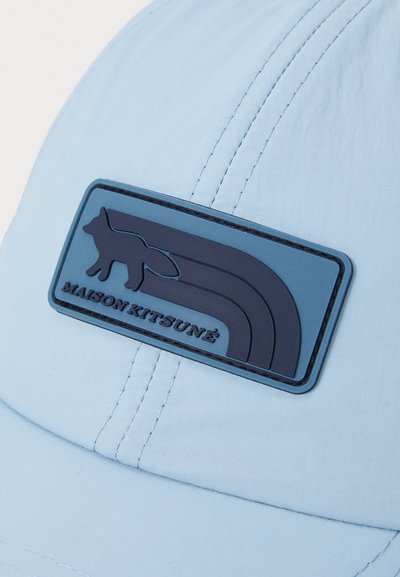 Casquette bleu clair en tissu au toucher lisse. Présente un patch en caoutchouc bleu foncé avec un motif de renard et des lignes courbées.