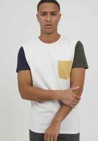 Vit t-shirt med korta ärmar i marinblått, grönt och gult. Har en gul ficka på vänster sida, gjord av mjukt bomullstyg.