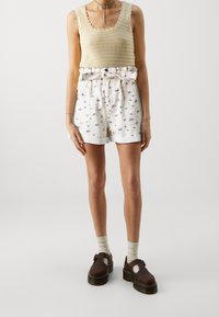 Débardeur en tricot beige avec des bords festonnés ; shorts blancs à motif floral bleu avec taille à nouer ; chaussures compensées marron foncé avec découpes.