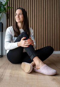 Felt slippers in light pink with a rubber sole, featuring a slip-on design and subtle stitching. Background has wooden slats and plants.