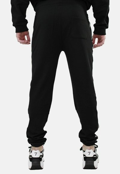 Pantaloni della tuta neri realizzati in un tessuto morbido, con un elastico in vita, design a gamba affusolata e una tasca posteriore. Abbinati a scarpe da ginnastica bianche.