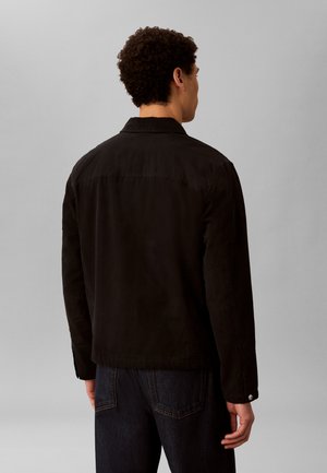 Mand med krøllet hår iført en sort jakke og mørke jeans, stående med ryggen mod en ensfarvet lys grå baggrund.