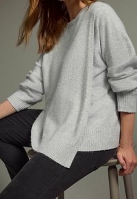 Pull gris clair à coupe décontractée, avec poignets et ourlet côtelés, présentant un tissu texturé et des fentes latérales, associé à un jean foncé.