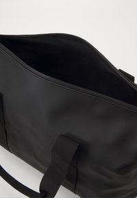 Sac de voyage étanche noir avec une fermeture éclair sur le dessus, en tissu résistant et doté de deux poignées de portage. Extérieur lisse avec un détail de branding subtil.