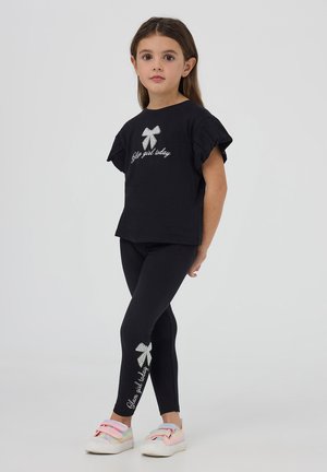 Maglietta nera a maniche corte con maniche arricciate e testo argentato. Leggings neri con design a fiocco e testo argentato. Sneakers luminose multicolori.