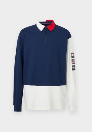 Μακρυμάνικο ράγκμπι πουκάμισο με σκούρο μπλε επάνω μέρος, λευκό κάτω μέρος, γιακά σε λευκό και κόκκινο, και επιθέματα στα μανίκια που περιλαμβάνουν τη σημαία των Ηνωμένων Πολιτειών και το λογότυπο της μάρκας.