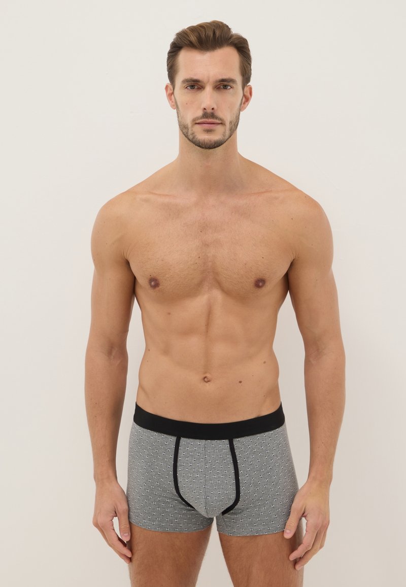 Homme torse nu aux cheveux bruns courts, portant un boxer gris à motifs avec une ceinture noire, debout devant un fond clair uni.