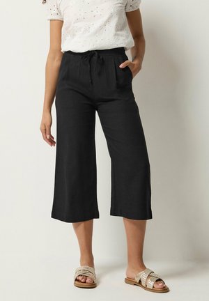 Mujer con pantalones negros anchos de pierna corta con cintura de cordón, blusa blanca de manga corta con ojales y sandalias planas tejidas, de pie junto a una pared.