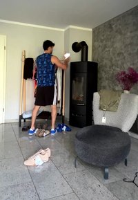 Άνδρας με αθλητική ενδυμασία επιλέγει ρούχα από ένα ξύλινο ράφι σε σαλόνι με μαύρη σόμπα και γκρι επενδεδυμένη καρέκλα.
