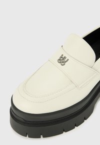 Mocassin en cuir blanc avec une finition brillante, doté d'une semelle plate noire et d'un logo métallique "HUGO" à l'avant. Design simpliste, couture minimale.
