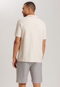 Camisa polo de manga corta de color beige con un cuello puntiagudo, combinada con pantalones cortos a rayas azules y blancas. Textura suave, diseño casual.
