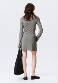 Rochie cu mâneci lungi, ajustată, din material verde măsliniu, ajungând până la mijlocul coapsei. Asortată cu o geantă de umăr texturată, neagră, cu butoane din argint.
