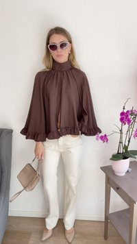 Top marrom de mangas sino com bainha de babados, combinado com calças brancas ajustadas e sapatos de salto bege claro. Segurando uma pequena bolsa bege.