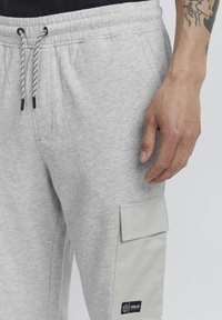 Man wearing light grey sweatpants with a drawstring waist, side pocket with flap, and tattooed forearm visible.