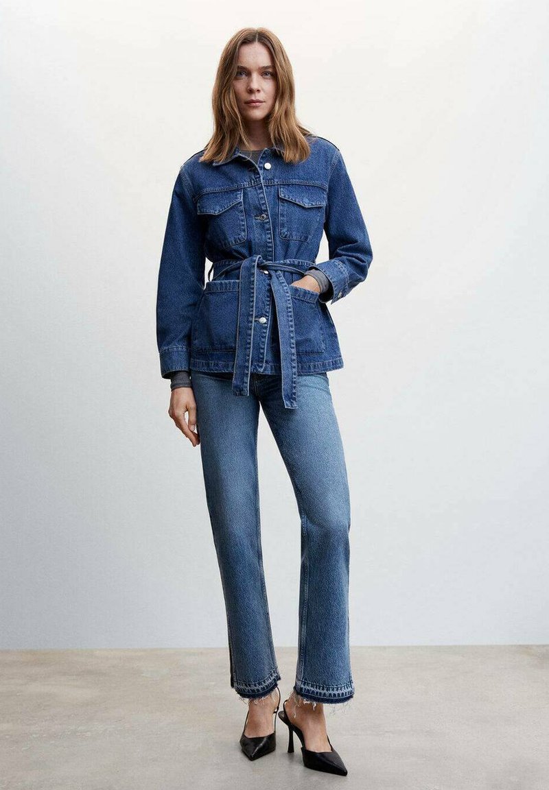 Veste en denim avec une taille ceinturée, poches poitrine et fermeture à boutons, associée à un jean évasé avec un ourlet effiloché et des talons foncés.