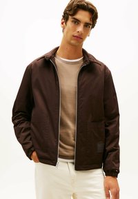 Brązowa kurtka na zamek z kołnierzem, z dwoma przednimi kieszeniami i elastycznymi mankietami, w połączeniu z beżowym swetrem i jasnymi spodniami.