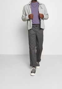 Homme portant une veste zippée grise clair, un t-shirt violet, un pantalon gris foncé, des baskets noires et blanches, se tenant devant un fond uni.