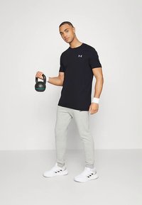 Muž v černém tričku a šedých teplácích drží kettlebell v pravé ruce a stojí na bílém pozadí.