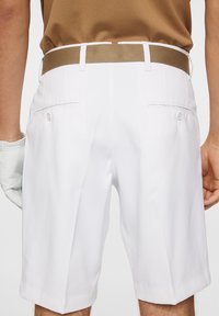 Białe szorty wykonane z gładkiego materiału, z brązowym paskiem i tylnymi kieszeniami zapinanymi na guziki. Zawierają rękę w jasnej rękawiczce.