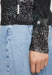 Zwarte langemouwen shirt met een gespikkeld wit en groen patroon, voorzien van een sterdetail, gecombineerd met lichtblauwe denim.