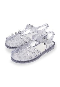 Sandalias de gel transparentes con un diseño de tiras, adornadas con tachuelas plateadas y acentos circulares. Cuenta con una correa en el tobillo con cierre de hebilla.