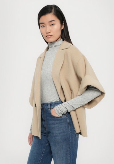 Beige outerwear with shawl sleeves, layered over a light gray turtleneck. Jeans with a high waist, featuring a button closure and side pockets.