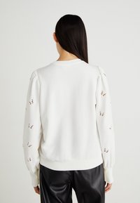 Sudadera blanca con mangas abullonadas que presenta patrones de mariposas recortados. Puños y dobladillo acanalados, textura suave y corte relajado.