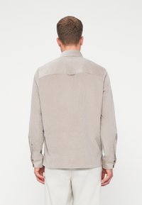 Camisa de botones color beige claro con una tela texturizada, mangas largas y un detalle de yugo en la parte de atrás. Presenta puños de botón individuales y un corte relajado.