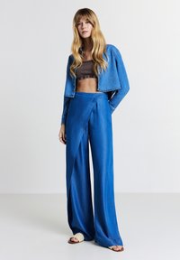 Donna con lunghi capelli biondi che indossa una giacca di jeans corta, un top corto marrone, pantaloni larghi blu e sandali beige aperti davanti, in piedi contro uno sfondo bianco.