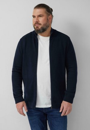 Mężczyzna z brodą i związaną z tyłu fryzurą, ubrany w granatowy sweter na zamek na białej koszulce i niebieskich dżinsach, patrzący w bok.