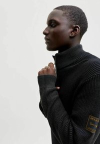 Maglione nero lavorato a maglia con collo alto, motivo strutturato e chiusura con cerniera. Include una toppa con logo sulla manica sinistra.