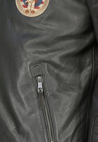 Schwarze Lederjacke mit strukturiertem Finish, einer Seitentasche mit Reißverschluss und einem gestickten Patch, der einen Adler darstellt.