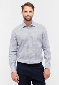 Camicia azzurro chiaro con un motivo geometrico, colletto button-down e maniche lunghe, abbinata a pantaloni scuri. Tessuto dalla consistenza liscia.