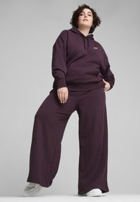 Felpa viola e pantaloni sportivi a gamba larga realizzati in tessuto morbido; presentano una tasca frontale e un dettaglio con logo, abbinati a sneakers bianche.