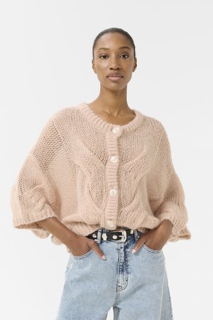 Cardigan en tricot rose doux avec un col rond, présentant un motif texturé, une fermeture à boutons et des manches amples. Assorti à un jean en denim clair.