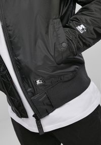 Giubbotto bomber nero con una texture liscia e lucida. Presenta polsini e orlo a coste, chiusura con zip e una piccola toppa con logo sulla tasca.