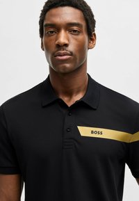 Sort polo shirt lavet af åndbart stof, med en gylden diagonale stribe og "BOSS"-logo på venstre brystområde. Design med tre-knap lukning.