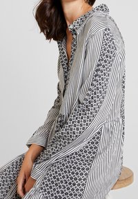 Robe noire et blanche avec des rayures verticales et des motifs géométriques, col boutonné et manches longues, confectionnée dans un tissu léger.