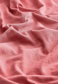 Tessuto color corallo rosa drappeggiato in morbide pieghe fluide. La texture appare leggera, liscia e leggermente sgualcita, mostrando un intreccio delicato.