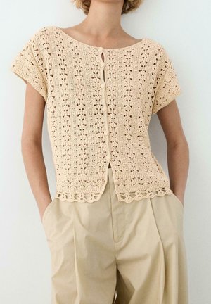 Kvinde iført en beige kortærmet hæklecardigan med knapper og beige bukser med højtaljede, hænderne i lommerne.