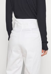 Witte broek met een brede tailleband en riemlussen. Heeft achterzakken en een gladde textuur. Gecombineerd met een zwarte geribbelde top met lange mouwen.