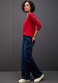 Czerwony sweter z dzianiny, szerokie ciemnoniebieskie dżinsy i białe trampki. Osoba stoi pewnie na tle o teksturze.