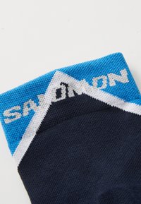 Calcetín azul marino con un diseño triangular en azul y blanco en la parte superior, que presenta "SALOMON" tejido en gris. Material texturizado con un acabado suave.