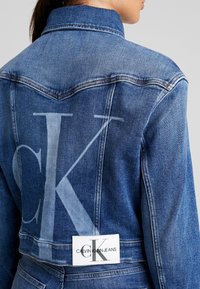 Žena v modré džínové bundě s velkým, vybledlým logem "CK" a bílou obdélníkovou etiketou Calvin Klein Jeans na zadní straně.