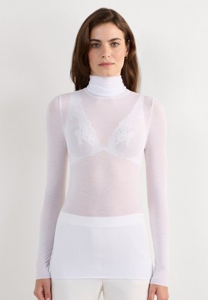 Mujer con cabello castaño que lleva una blusa blanca translúcida de manga larga con cuello alto y pantalones blancos sobre un fondo claro y sencillo.