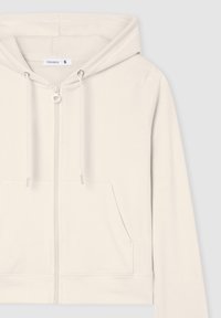 Felpa zip-up color crema in morbido tessuto. Presenta una tasca frontale, cappuccio con cordino e dettagli in metallo argentato. Design e texture semplici.