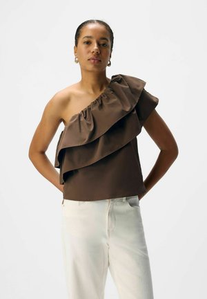 Haut marron à une épaule avec des volants superposés, présentant un col froncé. Associé à un pantalon ajusté de couleur claire.