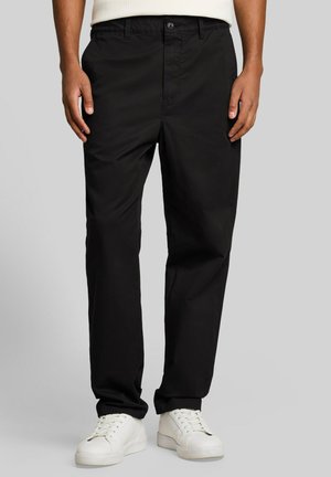 Person, die schwarze, gerade geschnittene Hosen und weiße Turnschuhe trägt, steht vor einem schlichten hellgrauen Hintergrund.