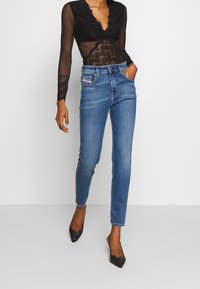 Vysoké modré džíny s úzkým střihem, které mají přední kapsu, doplněné o průsvitný černý krajkový top s dlouhými rukávy a černé podpatky.